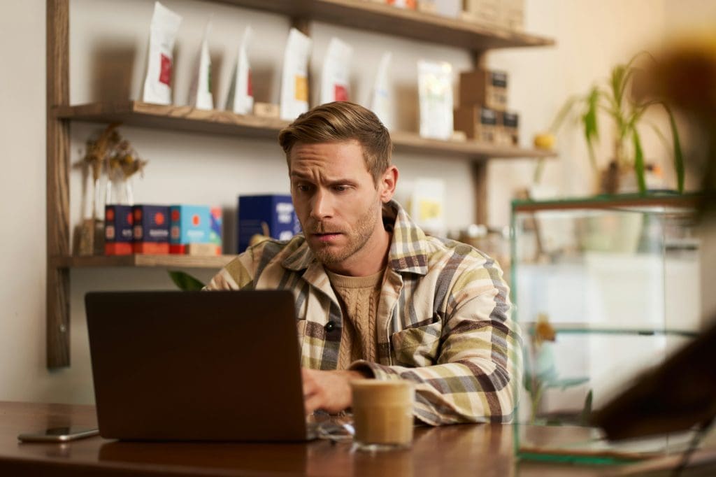 Image of young business owner, man sitting in cafe with laptop, looking confused at screen, digital